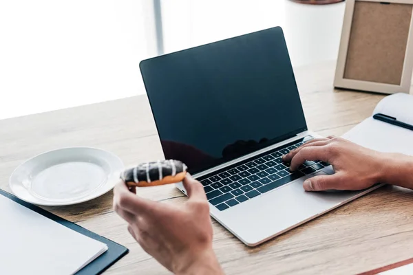 Tiro recortado de homem segurando donut e usando laptop à mesa com livro didático, área de transferência e moldura de fotos — Fotografia de Stock