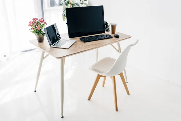 Интерьер рабочего места со стулом, цветами, кофе, канцелярскими принадлежностями, ноутбуком и компьютером на деревянном столе — стоковое фото