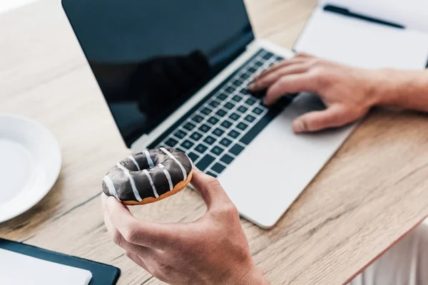 Visão parcial do homem segurando donut e usando laptop à mesa com livro didático e área de transferência — Fotografia de Stock