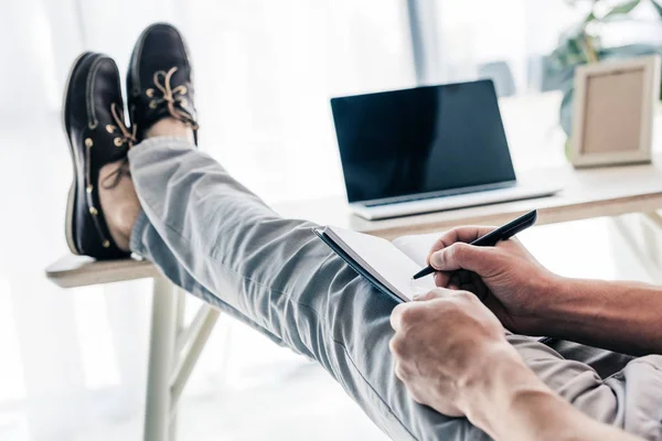 Cropped image of man writing in textbook and holding legs on table with laptop and photo frame — Stock Photo