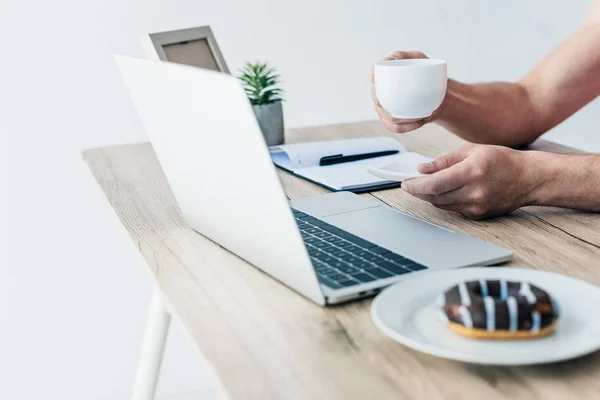 Visão parcial do homem segurando xícara de café na mesa com laptop, livro didático, moldura da foto, planta envasada e donut na placa — Fotografia de Stock