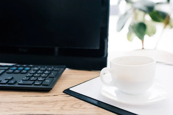 Vista de primer plano de la taza de café en el portapapeles vacío y el ordenador en la mesa de madera - foto de stock