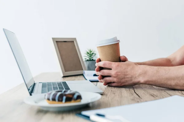 Hombre sosteniendo la taza de café en la mesa con el ordenador portátil, libro de texto, portapapeles, marco de fotos, planta en maceta y donut en el plato — Stock Photo