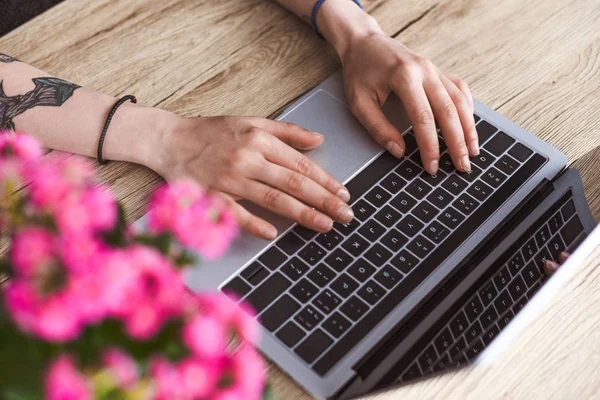 Vue partielle de la femme tatouée travaillant sur ordinateur portable à la table avec des fleurs — Photo de stock