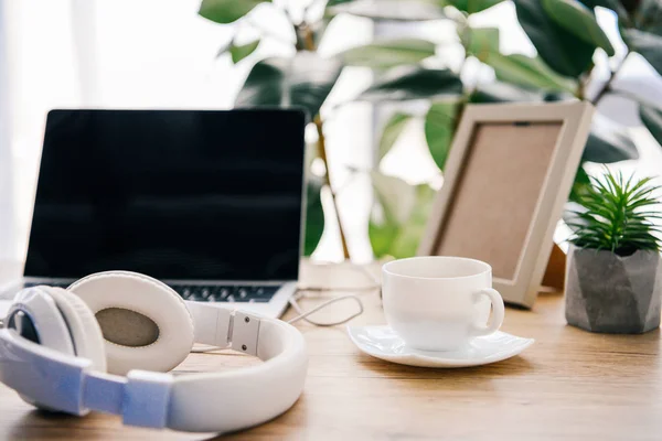 Vue rapprochée des écouteurs, ordinateur portable avec écran blanc, tasse à café, plante en pot et cadre photo — Photo de stock