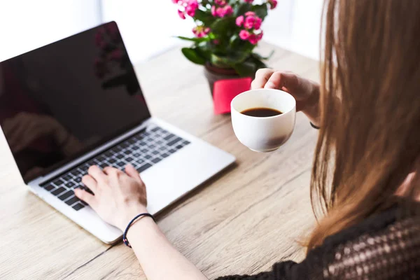 Vue partielle de la femme buvant du café et tapant sur ordinateur portable avec écran blanc à la table avec des fleurs — Photo de stock