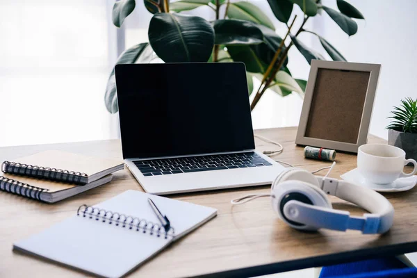 Vista de primer plano de la computadora portátil con pantalla en blanco en la mesa con auriculares, libros de texto, taza de café, marco de fotos, planta en macetas y rollo de dinero en efectivo - foto de stock