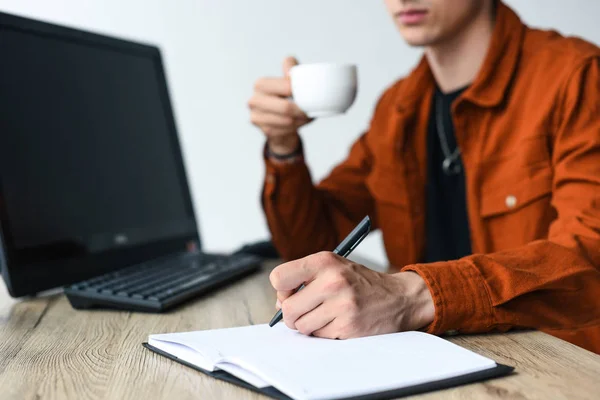 Частичный взгляд человека, пьющего кофе и пишущего в учебнике за столом с компьютером и клавиатурой — стоковое фото