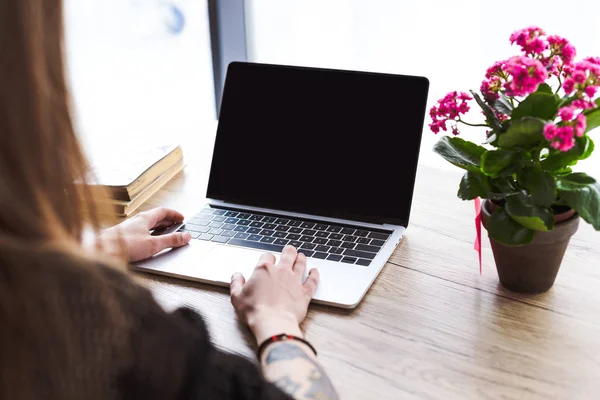 Image recadrée d'une femme tatouée tapant à la main sur un ordinateur portable à table avec des fleurs et des livres — Photo de stock