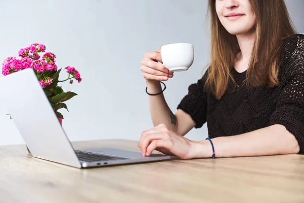 Частичный вид женщины, пьющей кофе и использующей ноутбук за столом с цветами — стоковое фото