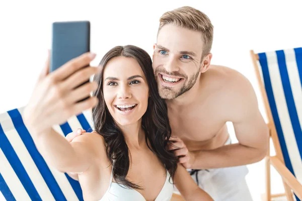 Hermosa pareja sonriente tomando selfie con teléfono inteligente en sillas de playa, aislado en blanco - foto de stock