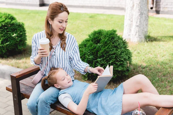 Criança sorrindo livro de leitura com a mãe por perto, enquanto descansando no banco juntos na rua — Fotografia de Stock