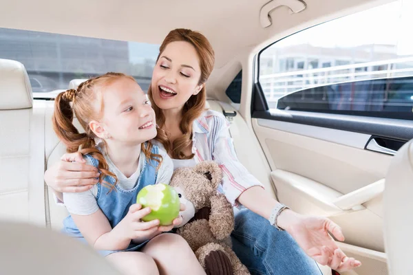 Happy mother and daughter with teddy bear and apple sitting in car — Stock Photo