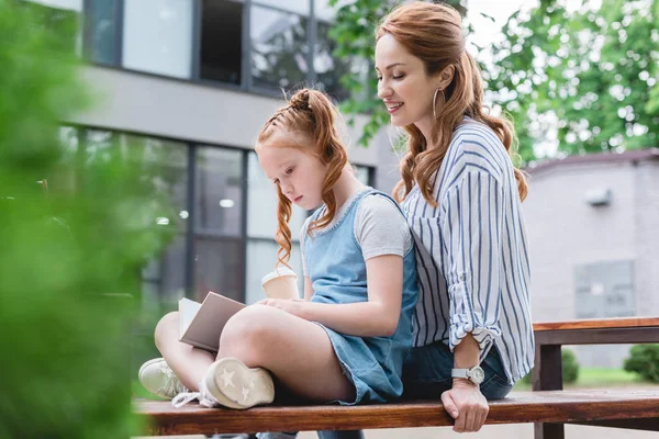 Kid reading book with mother near by while resting on bench together on street — Stock Photo