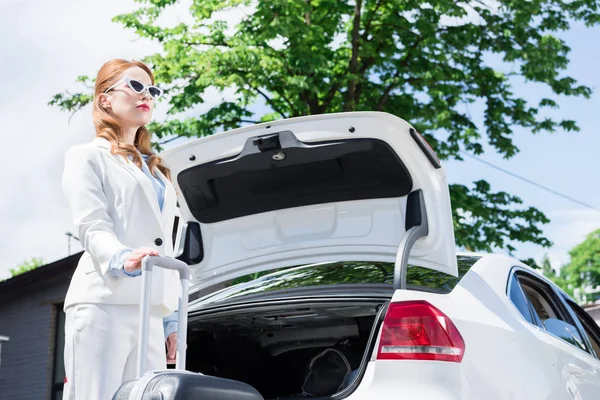 Mujer de negocios en traje blanco con maleta de pie en el coche - foto de stock
