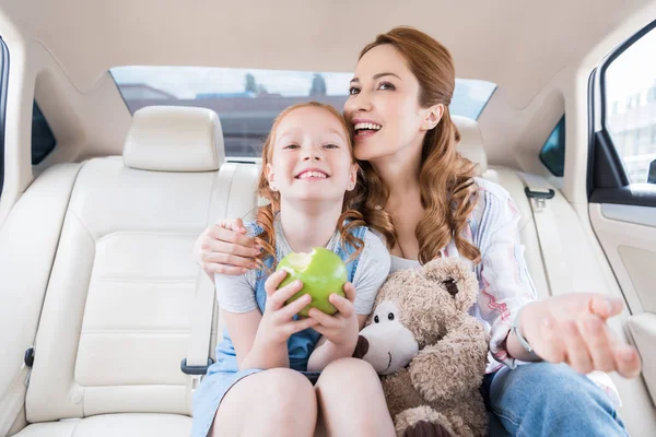 Ritratto di madre e figlia felici con orsacchiotto e mela seduti in macchina — Foto stock