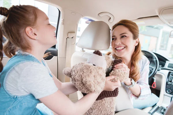 Seitenansicht der Tochter, die der lächelnden Mutter im Auto einen Teddybär gibt — Stockfoto