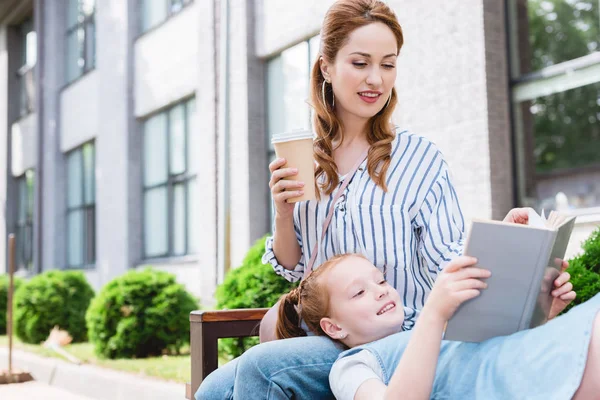 Kid reading book with mother near by while resting on bench together on street — Stock Photo