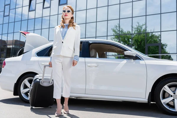 Деловая женщина в белом костюме и солнцезащитных очках с чемоданом, стоящим у машины — стоковое фото