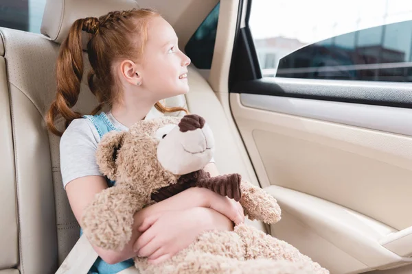 Portrait of cheerful kid hugging teddy bear and looking out car window while sitting in car — Stock Photo