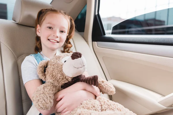 Retrato de niño alegre abrazando oso de peluche mientras está sentado en el coche - foto de stock