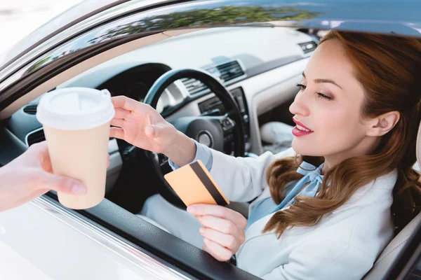 Businesswoman with credit card in hand taking coffee to go while sitting in car — Stock Photo