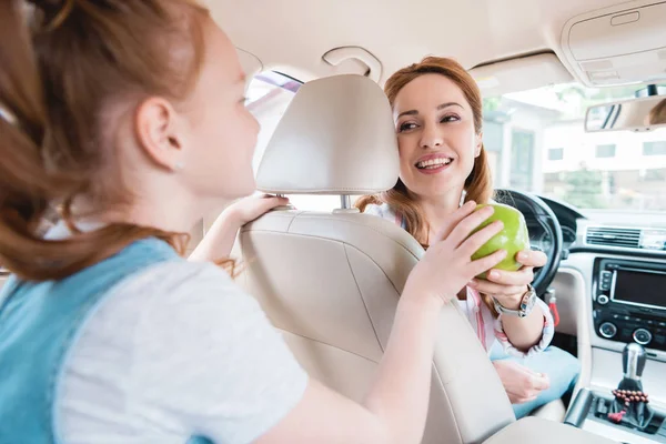 Vista lateral de la hija dando manzana fresca a la madre sonriente en el coche — Stock Photo