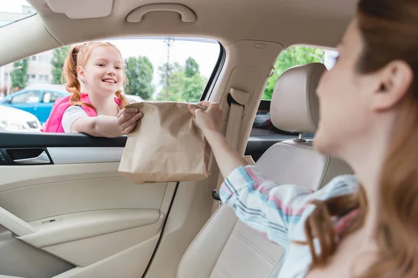 Vue partielle de la femme en voiture donnant paquet de papier avec de la nourriture à la fille — Photo de stock