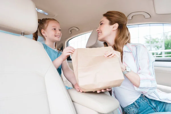 Mulher sorrindo dando pacote de papel com comida para filha no carro — Fotografia de Stock