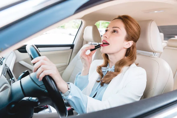 Vista lateral de la mujer de negocios aplicando lápiz labial mientras conduce el coche - foto de stock