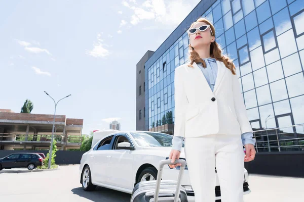 Vista de ángulo bajo de la mujer de negocios en traje blanco y gafas de sol con maleta de pie en el coche en la calle - foto de stock