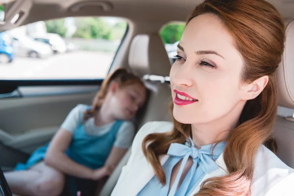 Избирательный фокус улыбающейся деловой женщины и спящей дочери на пассажирском сидении в автомобиле — стоковое фото