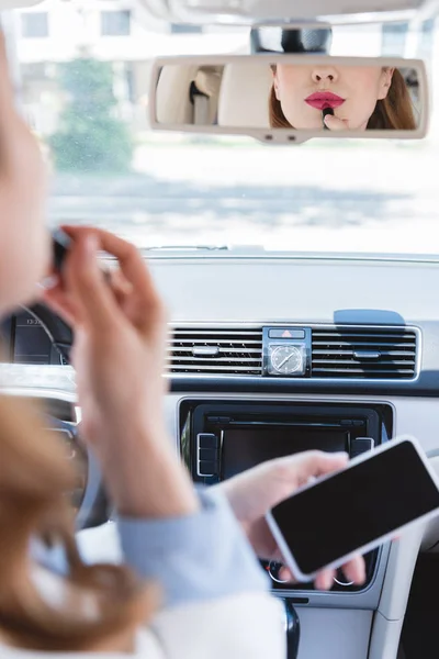 Partial view of businesswoman with smartphone in hand applying lipstick while driving car — Stock Photo