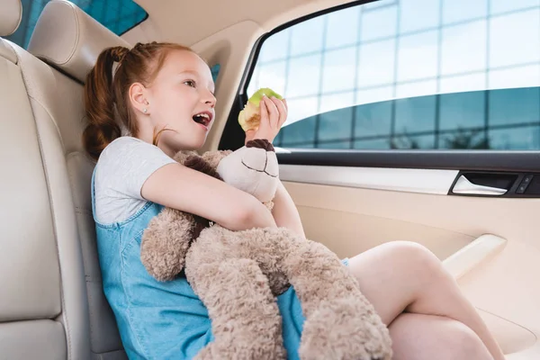 Vue latérale de l'enfant émotionnel avec ours en peluche et pomme fraîche en voiture — Photo de stock