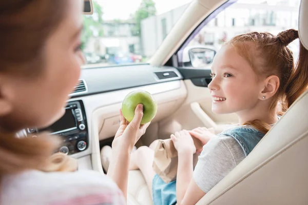 Vista parcial de la madre dando manzana a la hija sonriente en el coche - foto de stock