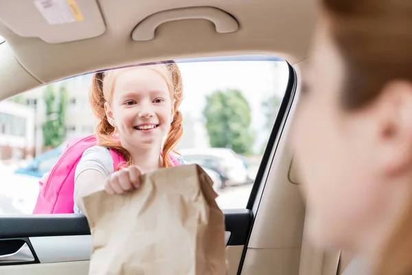 Частичный взгляд женщины в машине, дающей бумажный пакет с едой дочери — стоковое фото