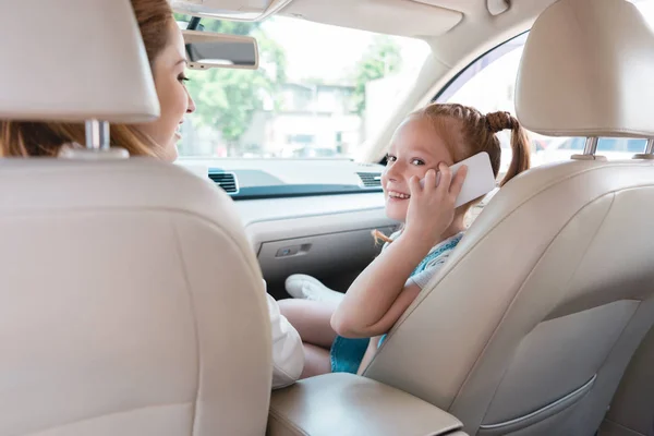 Seitenansicht eines lächelnden Kindes, das mit Mutter im Auto in der Nähe auf dem Smartphone spricht — Stockfoto
