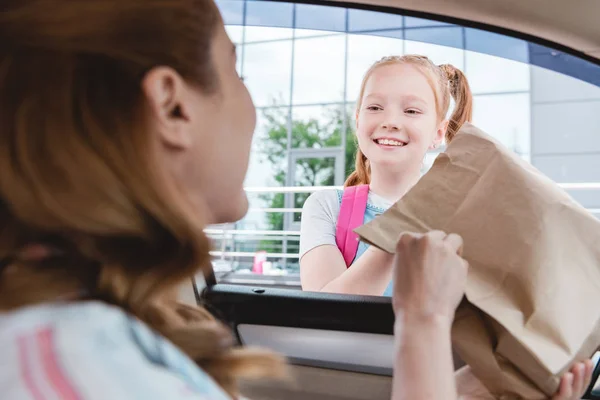 Selective focus of woman in car giving paper package with food to smiling daughter — Stock Photo