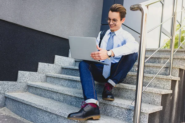 Low angle view of smiling young man in eyeglasses sitting on stairs and using laptop — Stock Photo