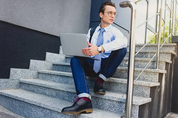 Bel giovane freelance con computer portatile seduto sulle scale e guardando altrove — Foto stock