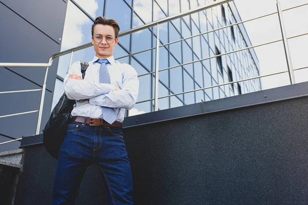 Tiefansicht eines hübschen jungen Geschäftsmannes, der mit verschränkten Armen vor einem Bürogebäude steht und in die Kamera blickt — Stockfoto