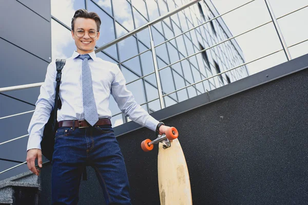 Low angle view of handsome young businessman in eyeglasses standing with longboard and smiling at camera — Stock Photo