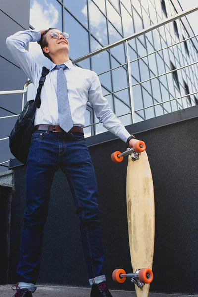 Smiling young businessman in eyeglasses standing with longboard and looking away — Stock Photo