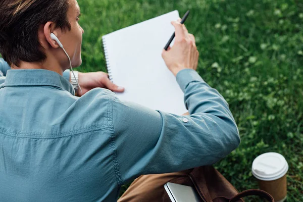 Tiro cortado de homem em fones de ouvido escrevendo em caderno em branco enquanto sentado na grama — Fotografia de Stock