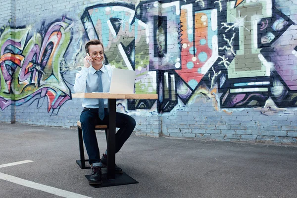 Smiling young freelancer in eyeglasses talking by smartphone and using laptop while sitting on street — Stock Photo