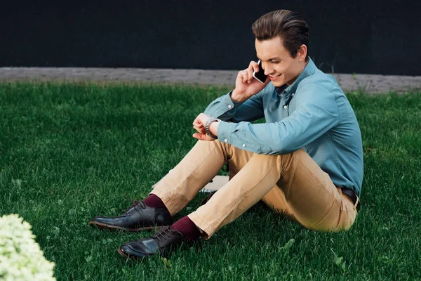 Souriant jeune homme parlant par smartphone et vérifier montre-bracelet tout en étant assis sur l'herbe — Photo de stock