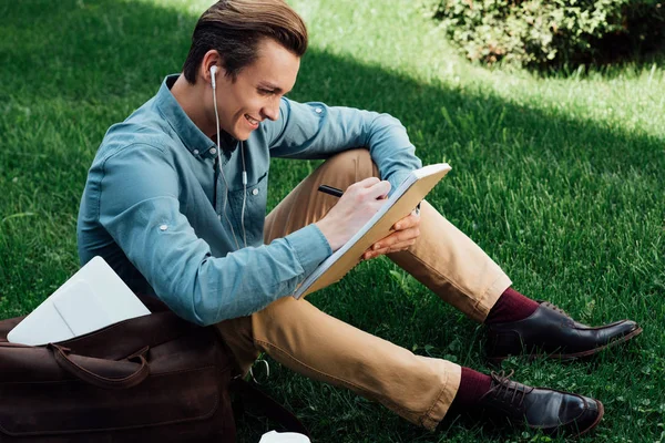 Vue grand angle du jeune homme souriant dans les écouteurs prenant des notes dans le bloc-notes — Photo de stock