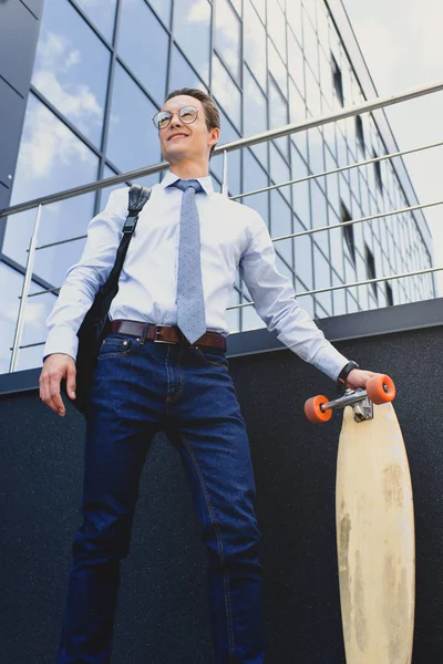 Low angle view of smiling young businessman in eyeglasses standing with longboard and looking away — Stock Photo