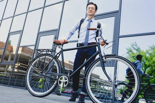 Low angle view of handsome young businessman in eyeglasses standing with bicycle near modern building — Stock Photo