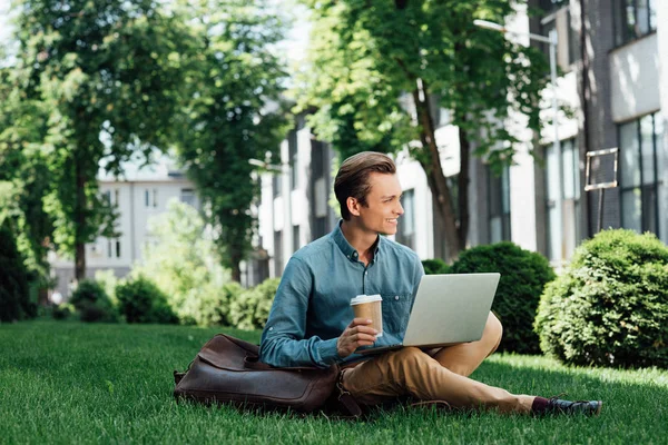Sonriente joven freelancer con café para ir sentado en la hierba y el uso de la computadora portátil - foto de stock